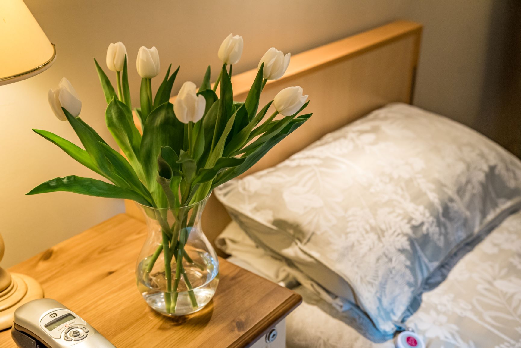 A resident's bedside table with a vase of flowers and fresh pillow.